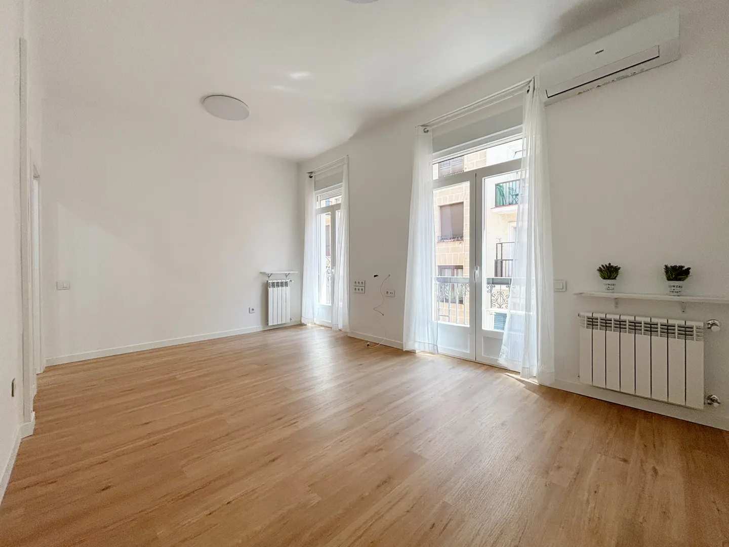 Bright, empty room with wood floors, white walls, and two sets of glass doors with sheer curtains. Radiators and AC unit are visible.