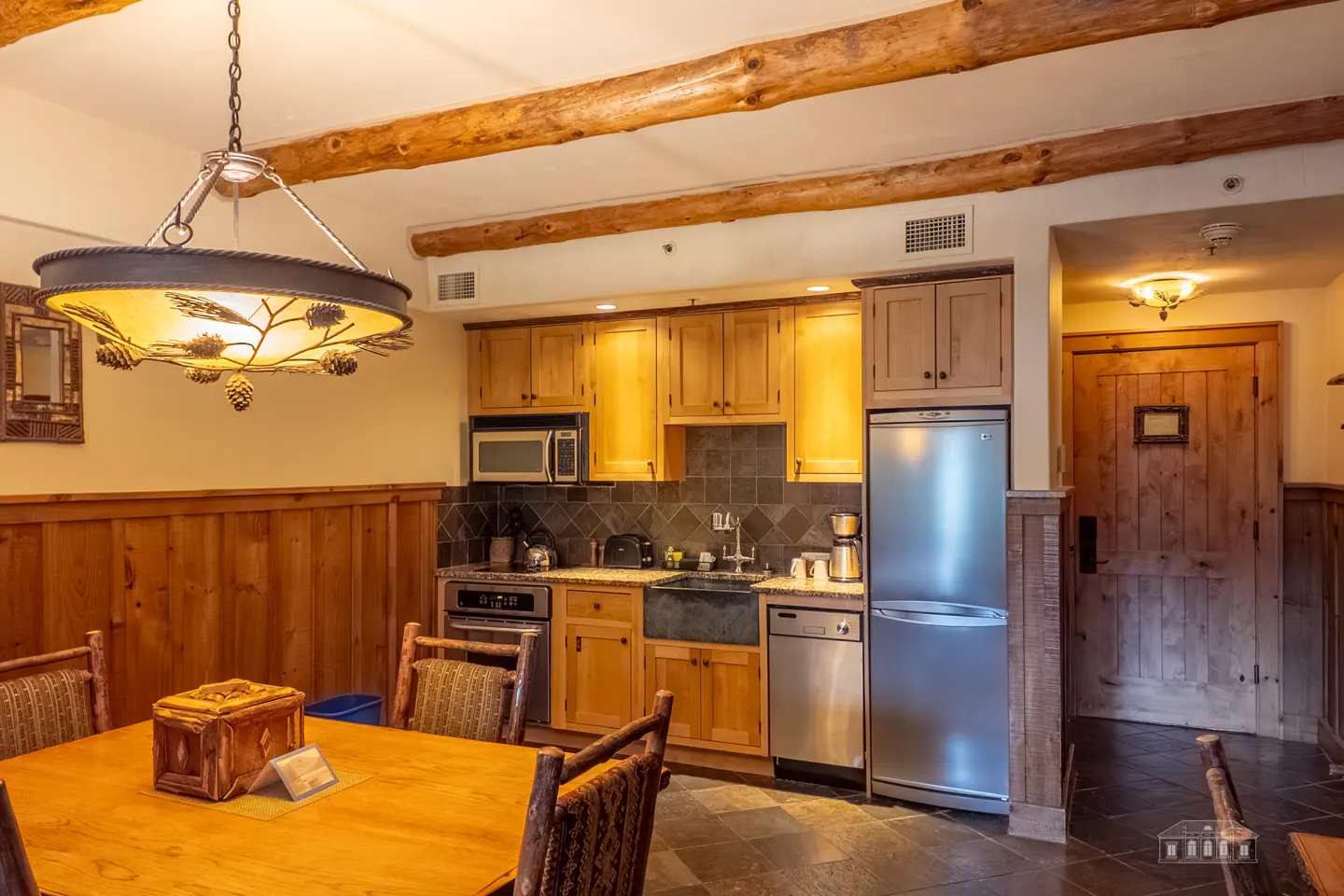 A warm-toned kitchen with wood cabinets, stainless steel appliances, and a dining table with wooden chairs. A rustic chandelier hangs above.