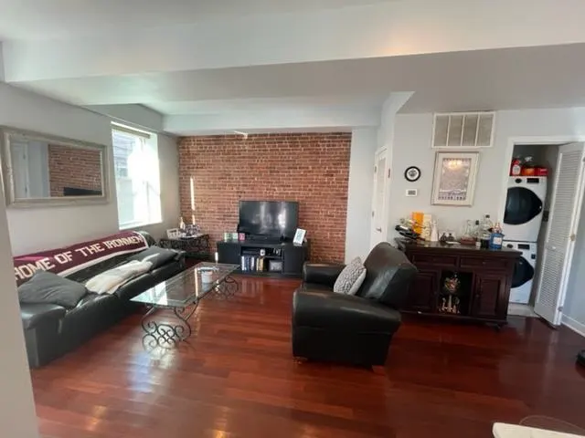 Living room with hardwood floors, brick accent wall, black leather sofa and chair, glass coffee table, and TV. Washer and dryer in closet.