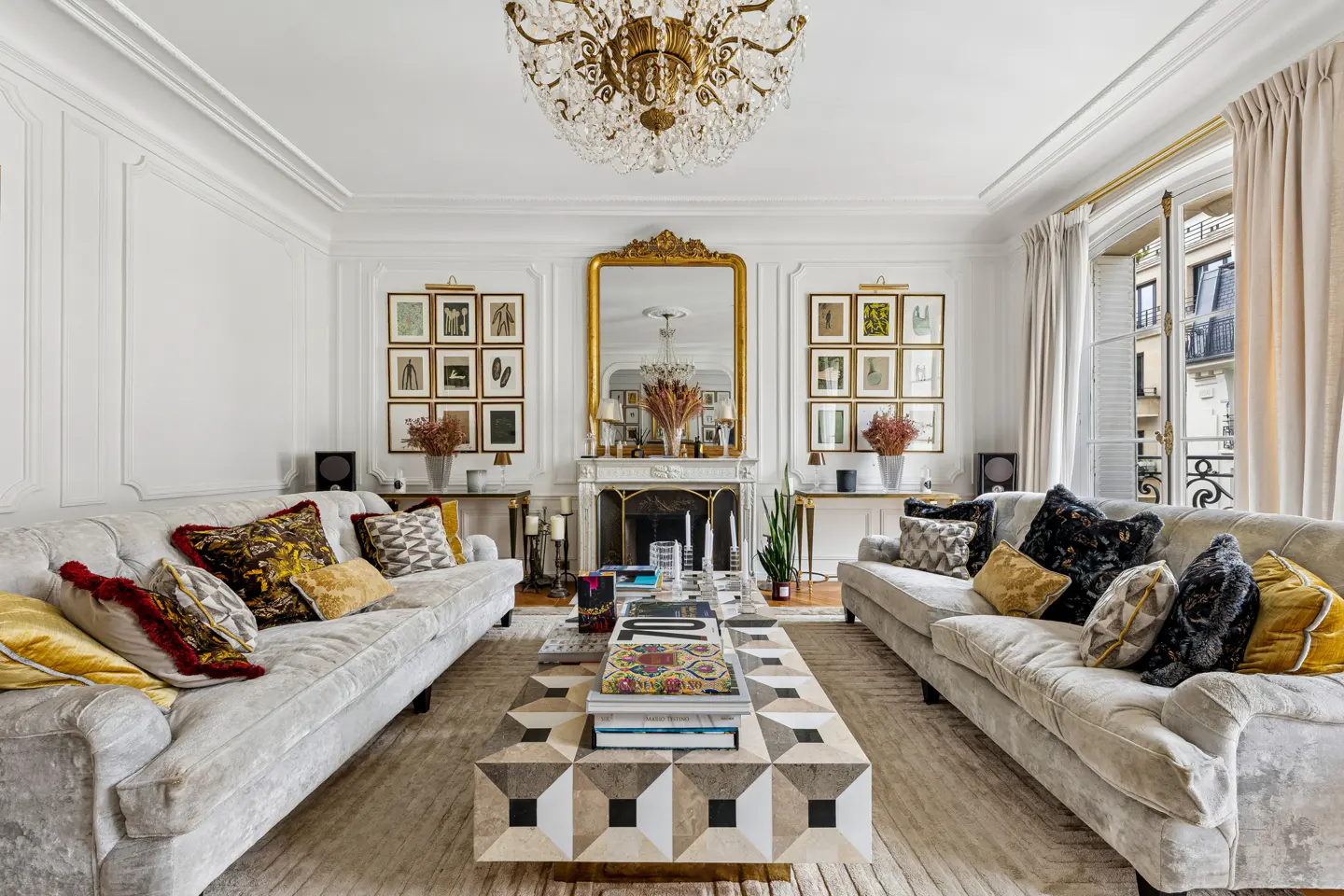 Elegant living room with two sofas facing each other, a patterned coffee table, and a gold-framed mirror above a fireplace. A chandelier hangs from the ceiling.