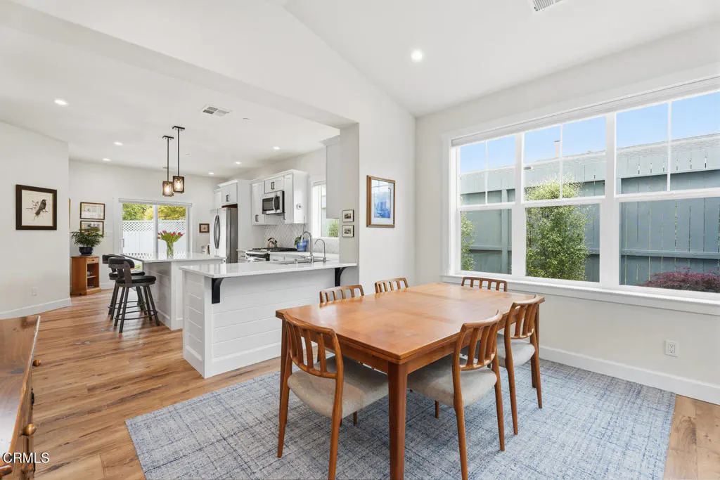 Bright dining area with a wooden table and chairs on a blue rug, opening into a modern white kitchen with an island.