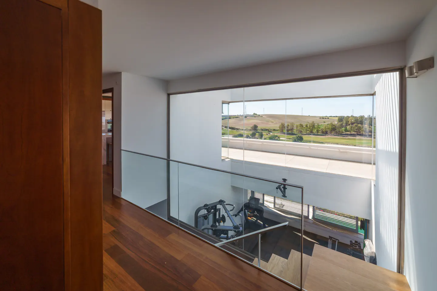 Hallway with wood floors and glass railing overlooking a gym. A large window shows a green landscape.