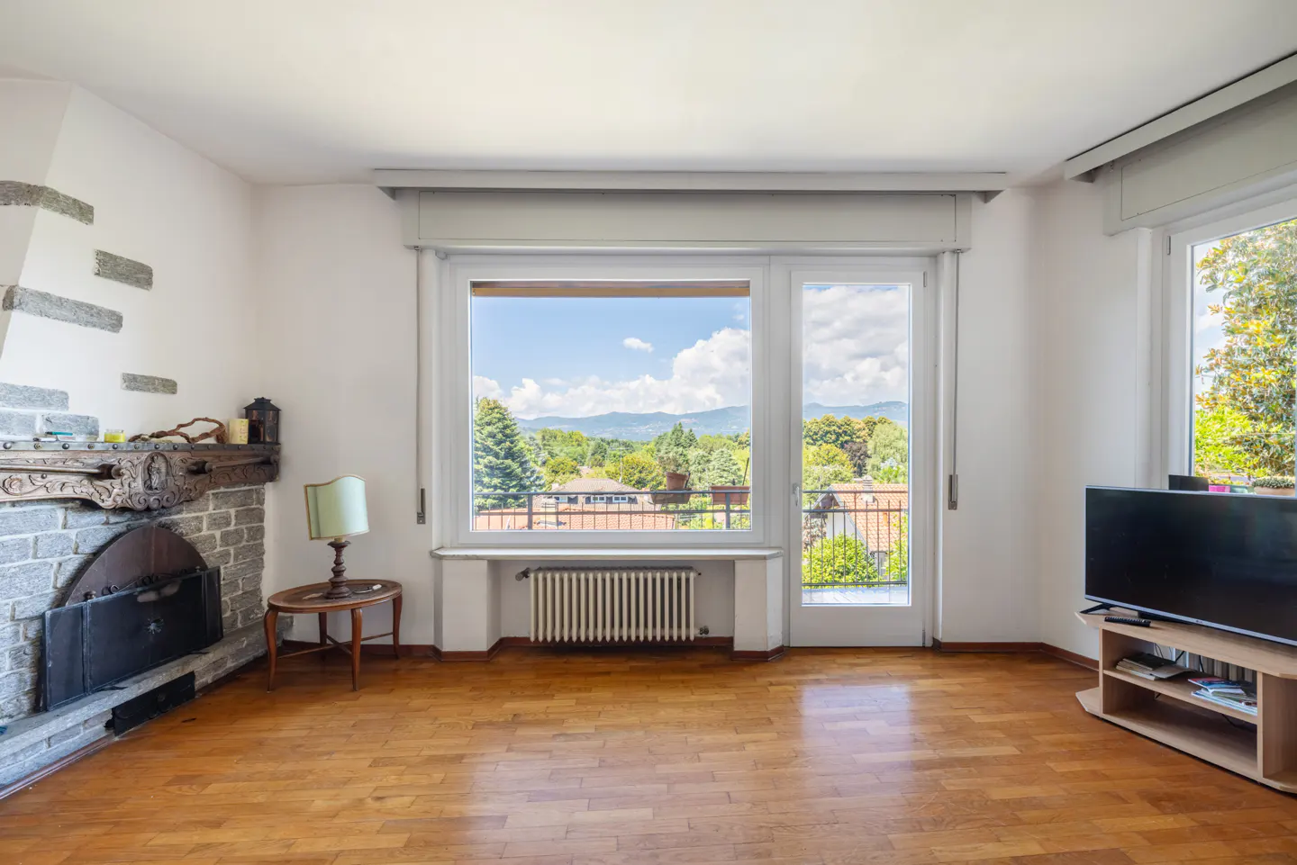 Bright living room with hardwood floors, stone fireplace, and a large window with a view of trees and mountains.