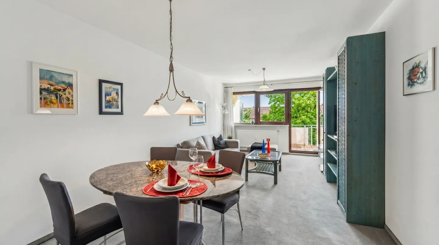 Dining area with a round marble table, four chairs, and a living room with a sofa and balcony view in the background.