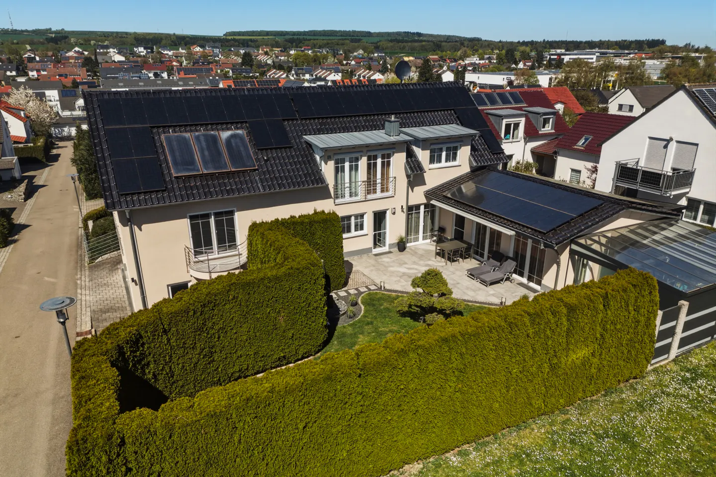 Aerial view of a beige two-story house with solar panels, a green hedge, and a patio with furniture.