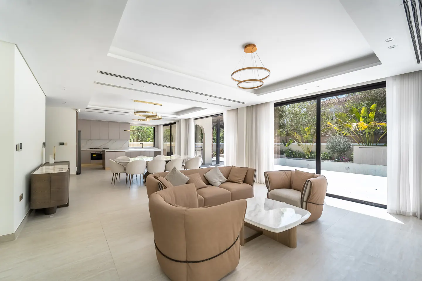 Bright, open-plan living space with tan sofa and chairs, marble coffee table, and floor-to-ceiling windows overlooking a lush garden.