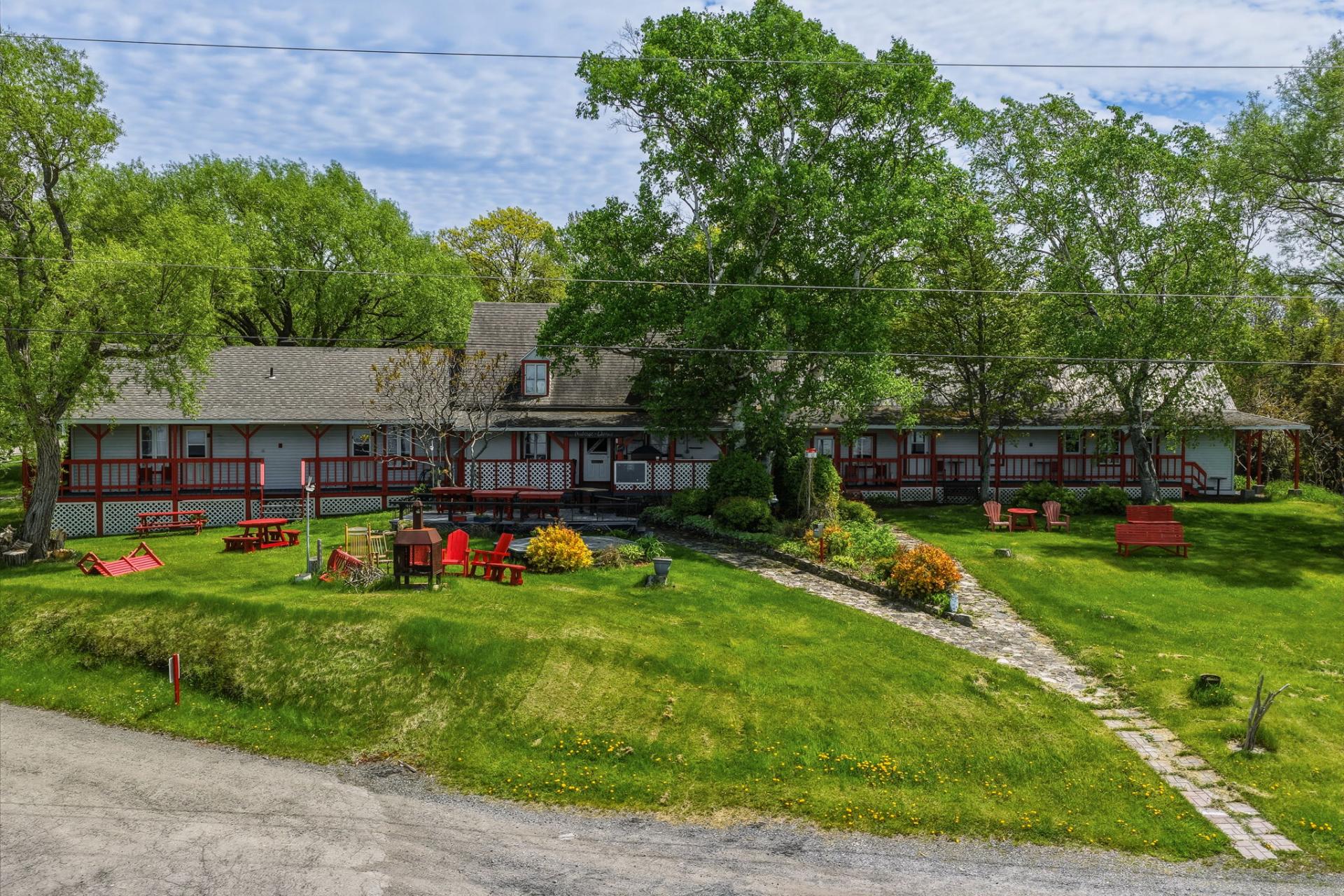 A long, grey building with red trim and a wrap-around deck sits on a lush green lawn with red outdoor furniture and trees. [AI-generated text]