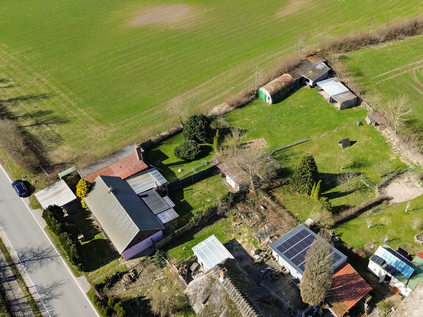 Aerial view of a rural property with a house, outbuildings, solar panels, and a large green field. A car is parked on the road.