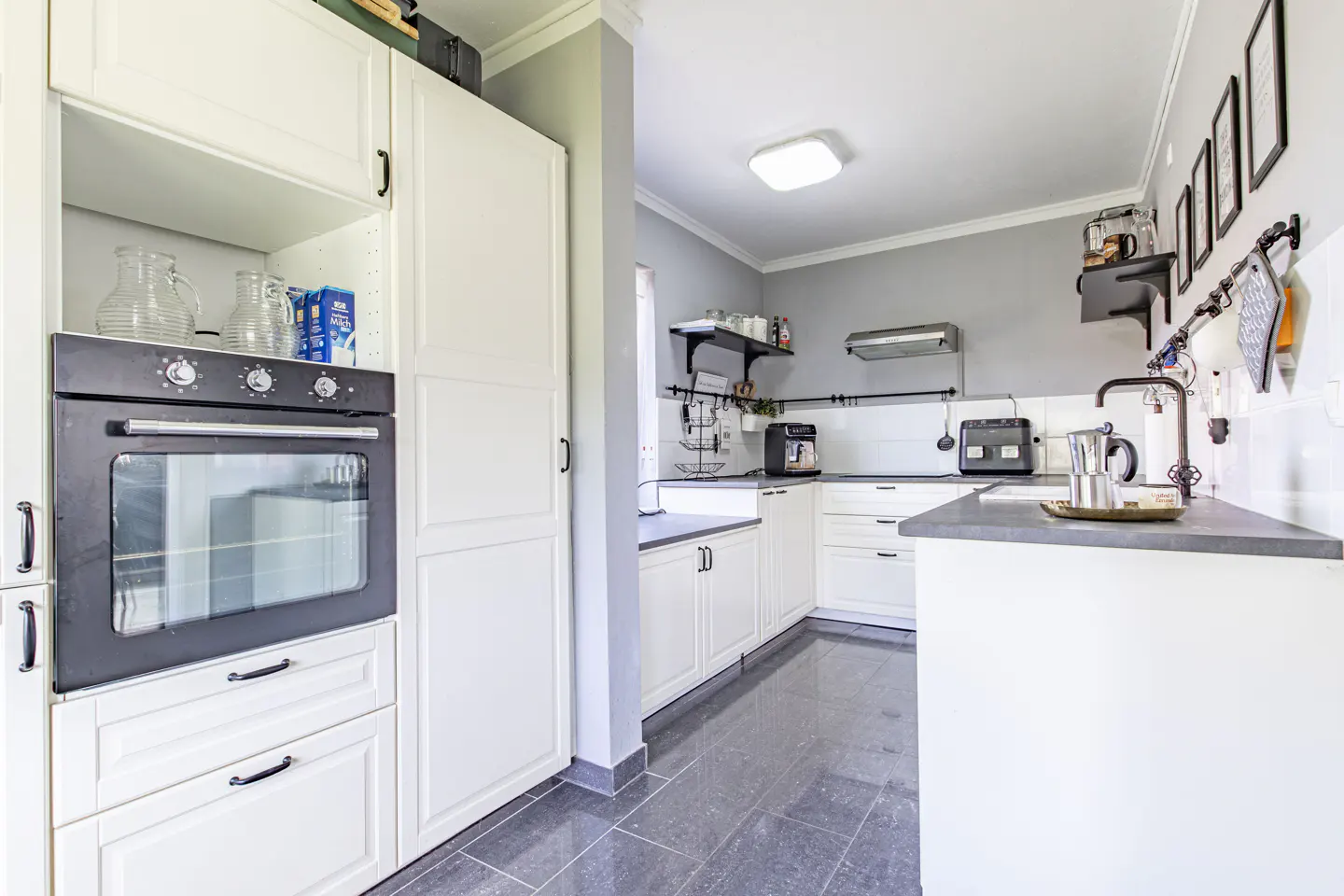 A bright kitchen with white cabinets, gray walls, and dark gray tile flooring. An oven is built into the cabinets on the left.