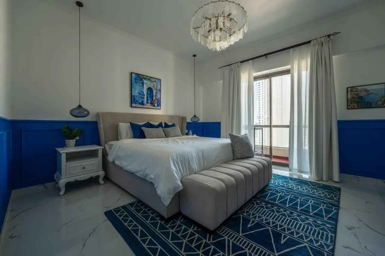 A bedroom with white walls, blue wainscoting, a king bed, and a blue patterned rug. A chandelier hangs from the ceiling.