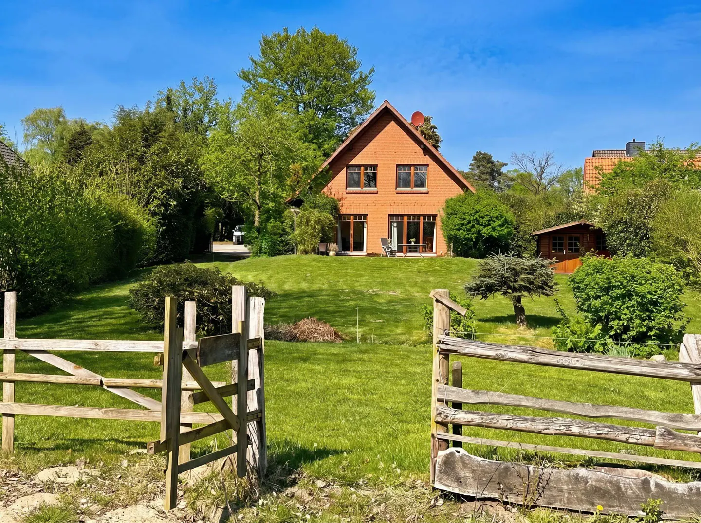 A brick house sits on a green lawn, viewed through an open wooden gate under a blue sky.