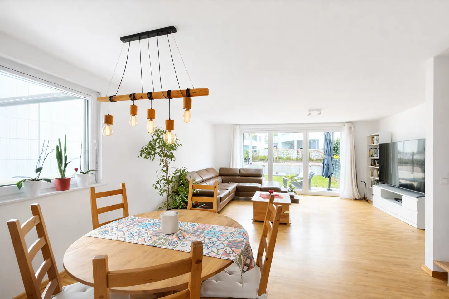 Bright, open-concept living space with a round wooden table, chairs, and a modern light fixture. A brown leather sofa is visible in the background.