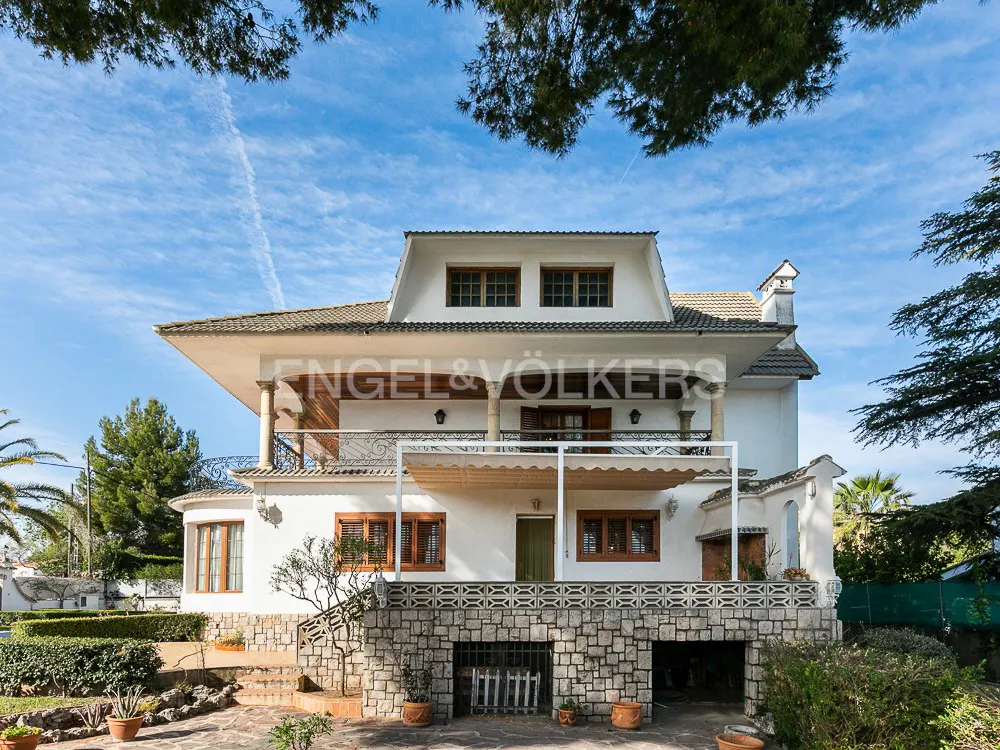 Two-story white house with a stone foundation, brown trim, and a balcony, under a blue sky with trees.