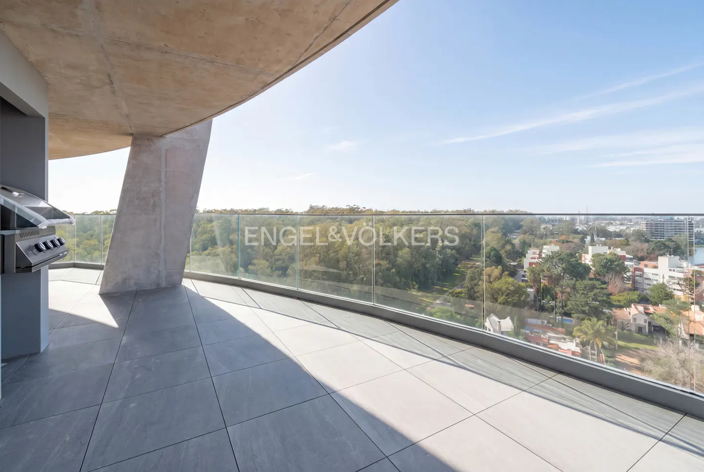 Spacious modern balcony with gray tiled flooring, a built-in grill, and a glass railing overlooking a lush green landscape.