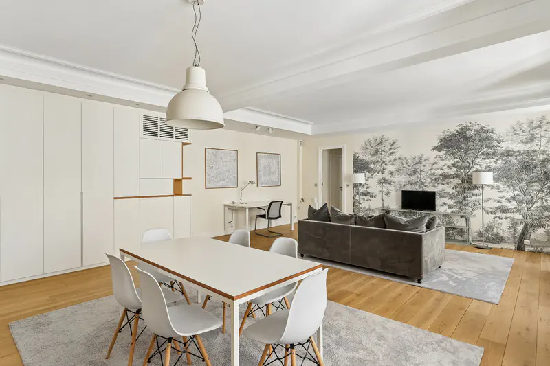 Bright, open-concept apartment with a white dining table, gray sofa, and a black and white forest mural. Hardwood floors and a large white pendant light.