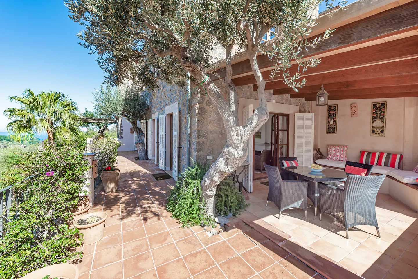 Outdoor patio with a dining table, chairs, and built-in bench seating. A large tree provides shade. The patio has terracotta tile flooring.