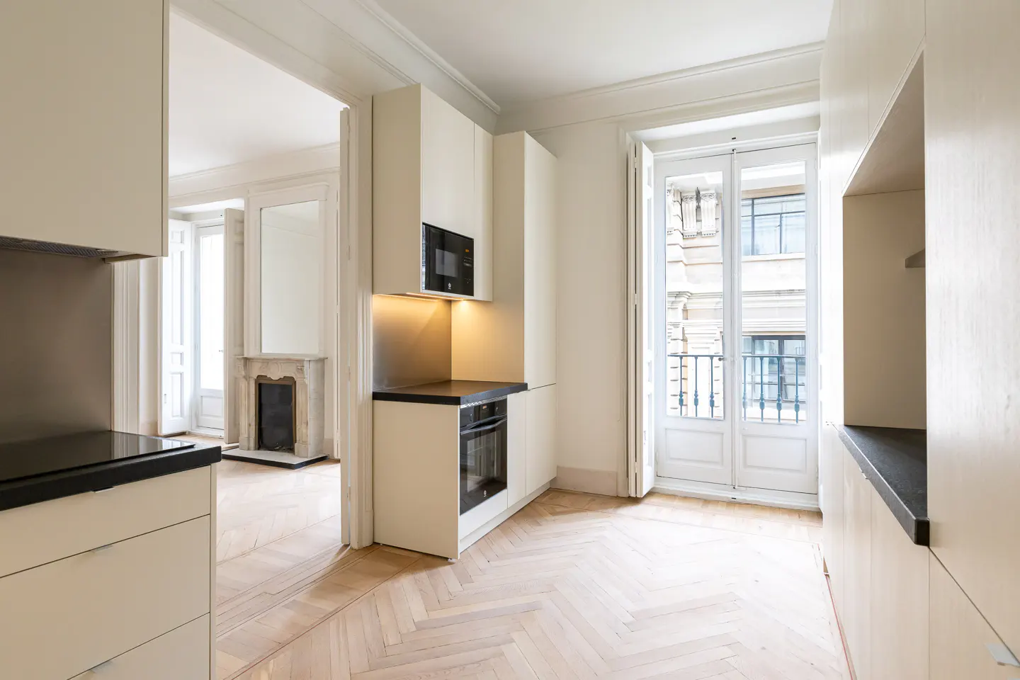 Bright, modern kitchen with white cabinets, black countertops, and herringbone wood floors. Balcony doors open to a city view.