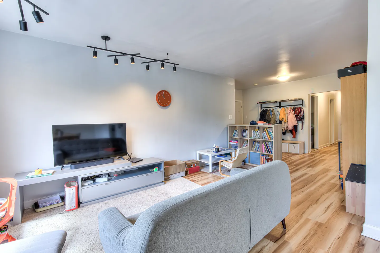 Living room with gray sofa, TV on a gray stand, and a bookshelf. Wood floors lead to a hallway with coats hanging on a rack.