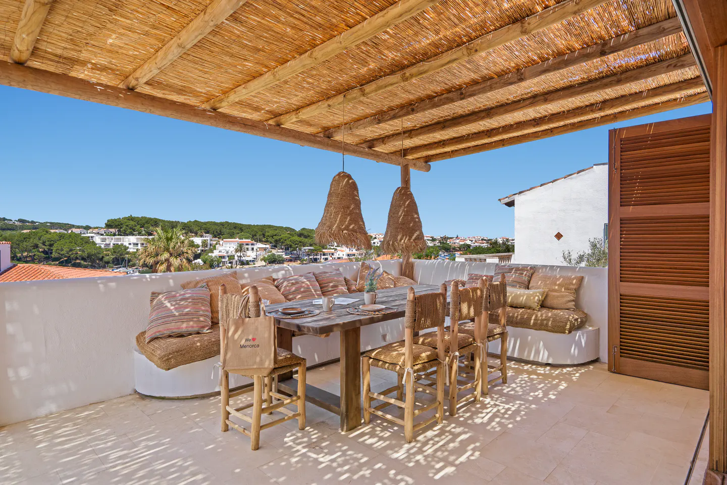 Outdoor dining area with a thatched roof, wooden table and chairs, and built-in seating with cushions, overlooking a town.