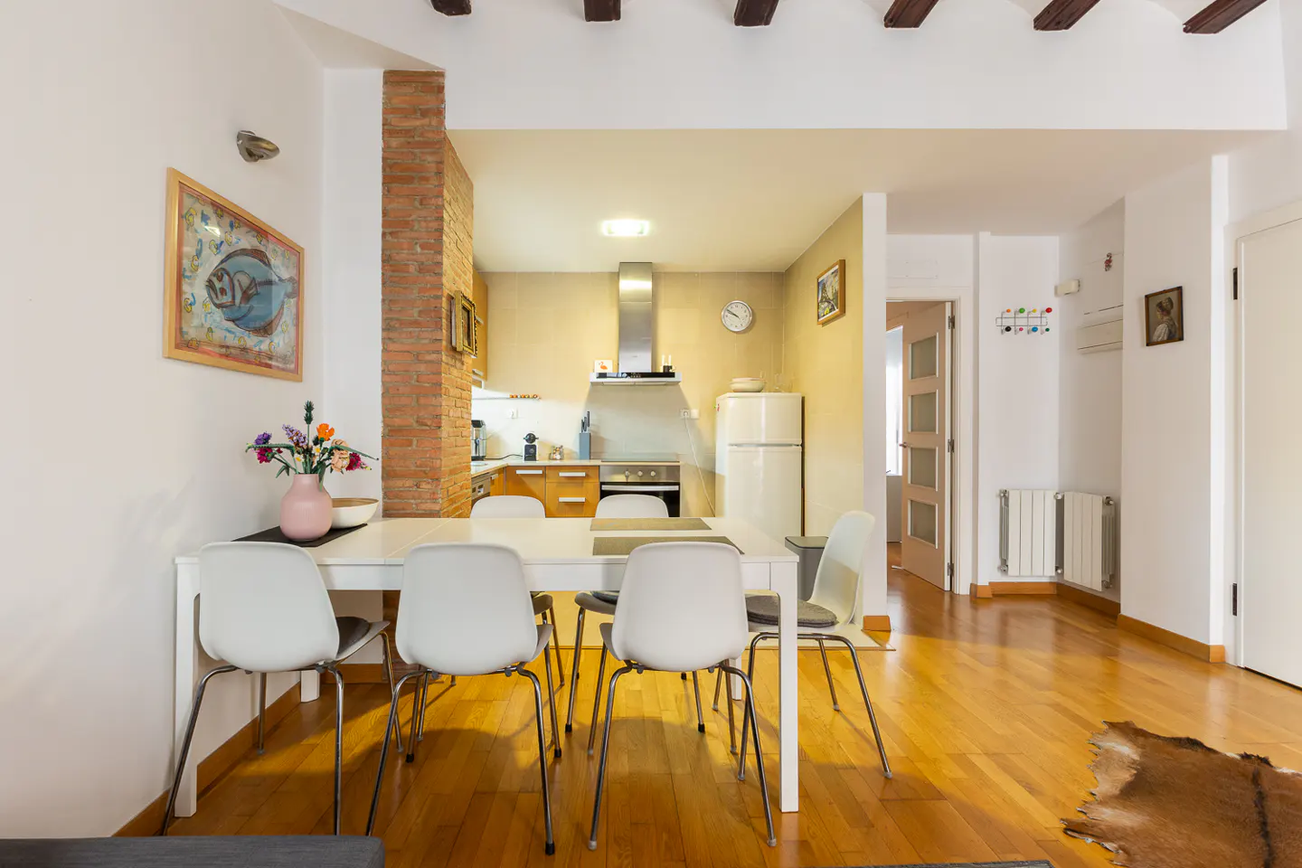 Bright, open-concept living space with a white dining table, chairs, and a view into the kitchen with wood cabinets and stainless steel appliances.