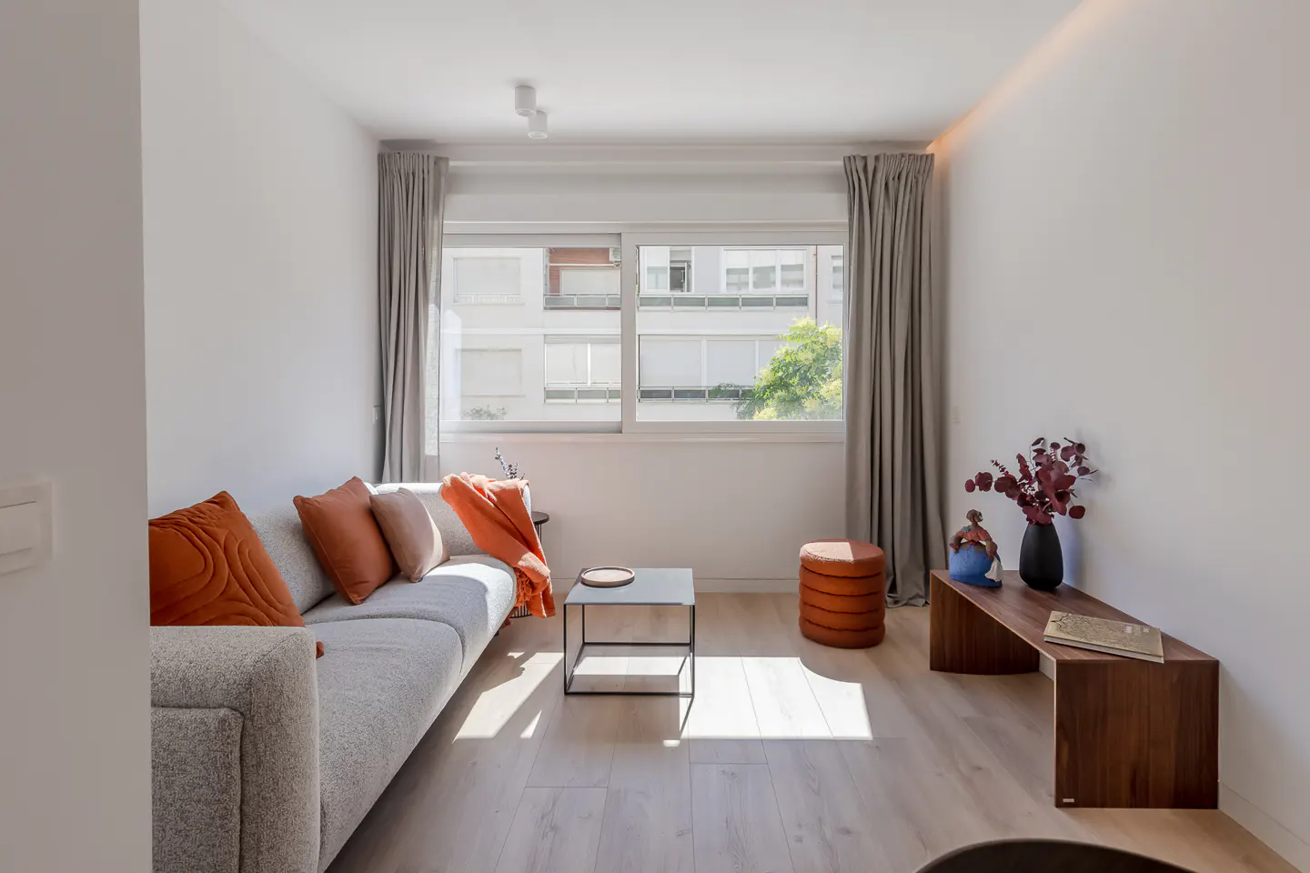 Bright living room with a gray sofa, orange pillows, a black table, and a wooden bench with a vase.