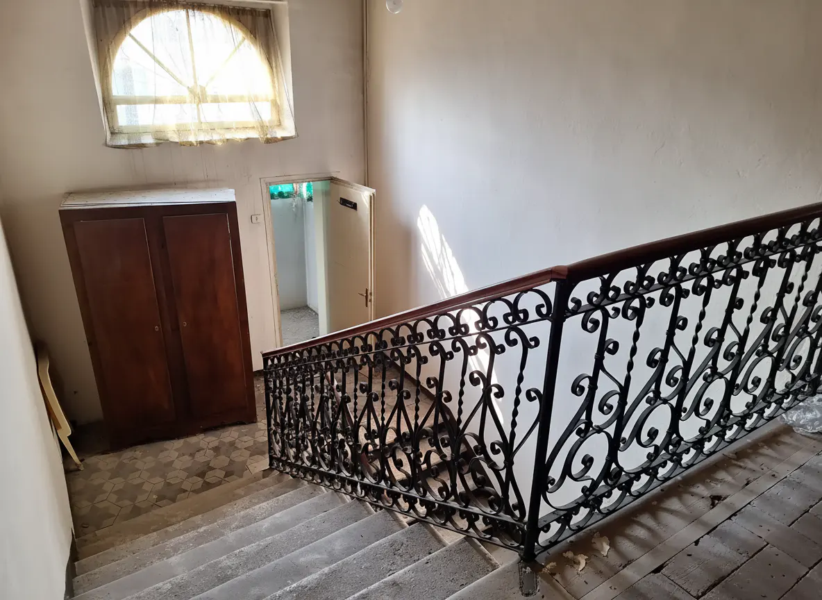 Interior view of a stairwell with a decorative black iron railing and a wooden handrail. A window and a brown wardrobe are visible.