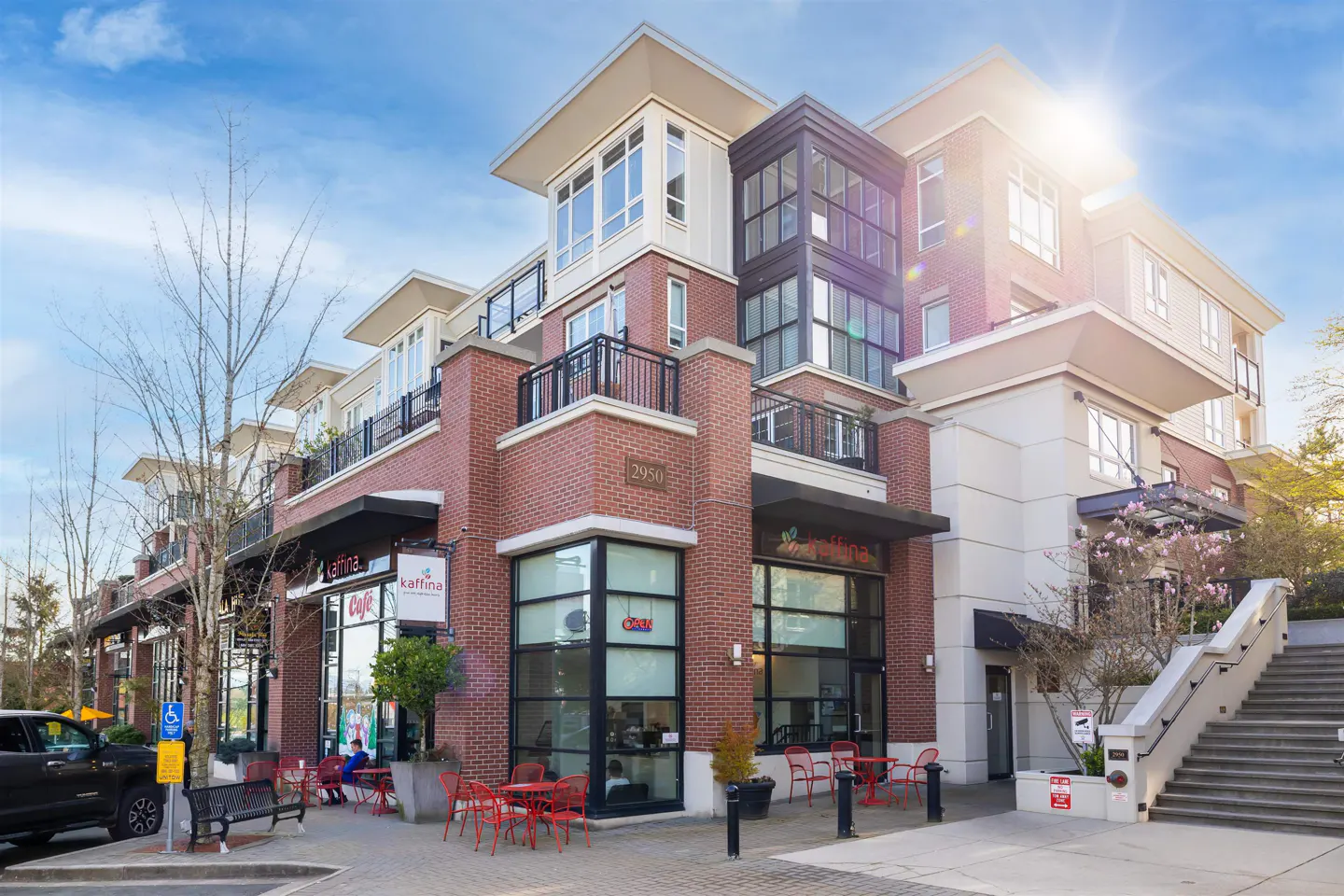 Exterior of a mixed-use building with a brick facade and a "Kaffina" cafe on the ground floor. Red chairs and tables are outside.