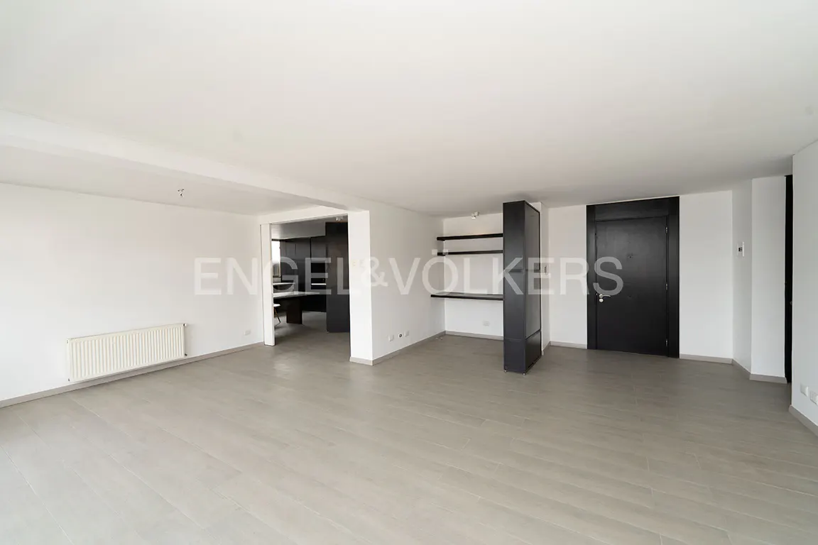 Bright, empty living room with light wood floors, white walls, black shelving, and a glimpse of a dark kitchen.