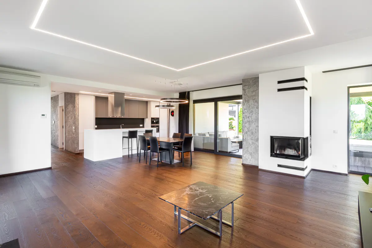 Open-concept living space with dark wood floors, white walls, and recessed lighting. Kitchen, dining area, and fireplace are visible.
