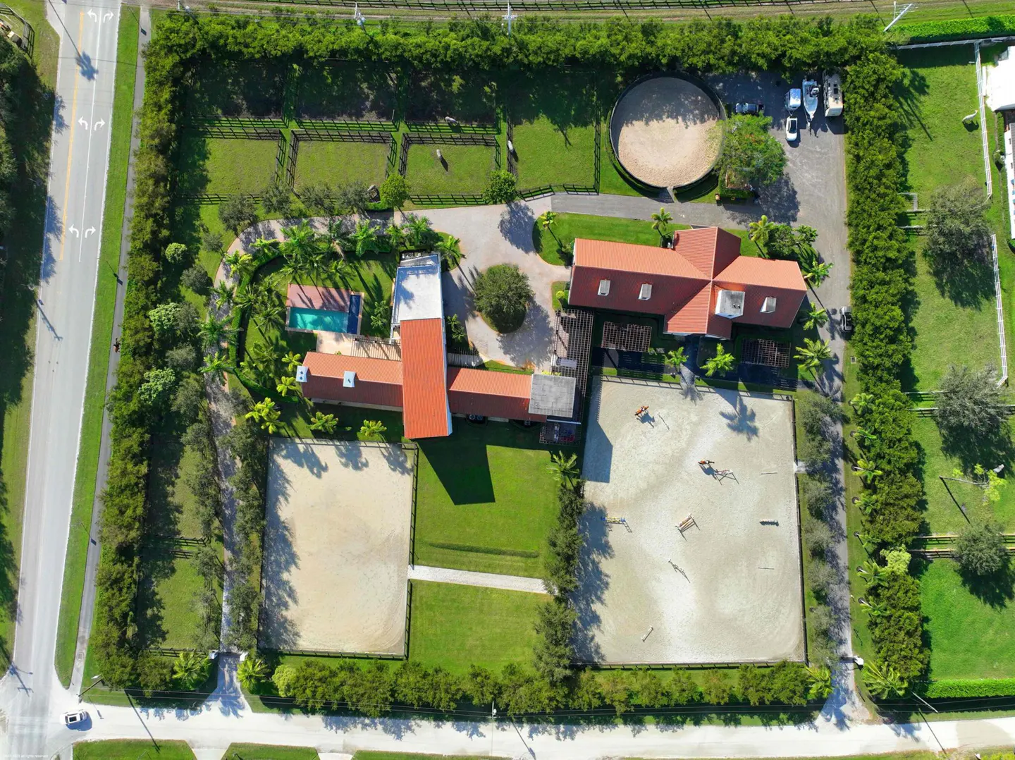 Aerial view of a horse farm with a red-roofed house, paddocks, riding arena, pool, and green landscaping.