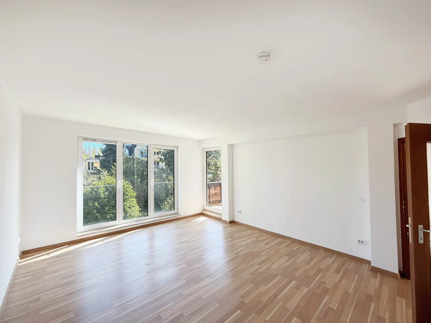 Bright, empty room with wood floors, white walls, and large windows showing trees and a building. A brown door is partially open.