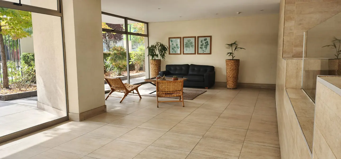 A bright lobby with tile floors, a black sofa, two wicker chairs, and large windows overlooking greenery.