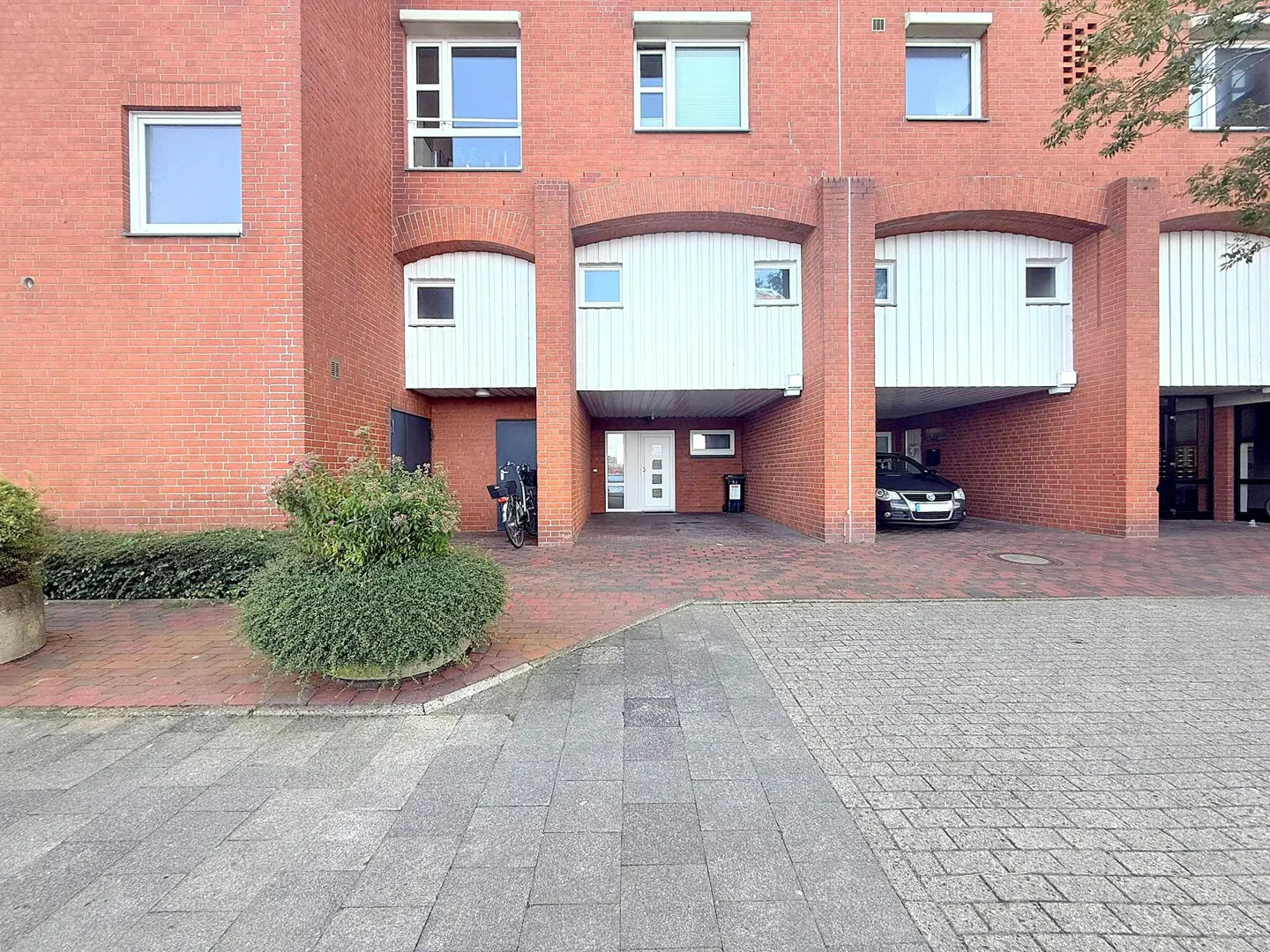Brick apartment building with carports. A black car is parked in one. A bush is in front.