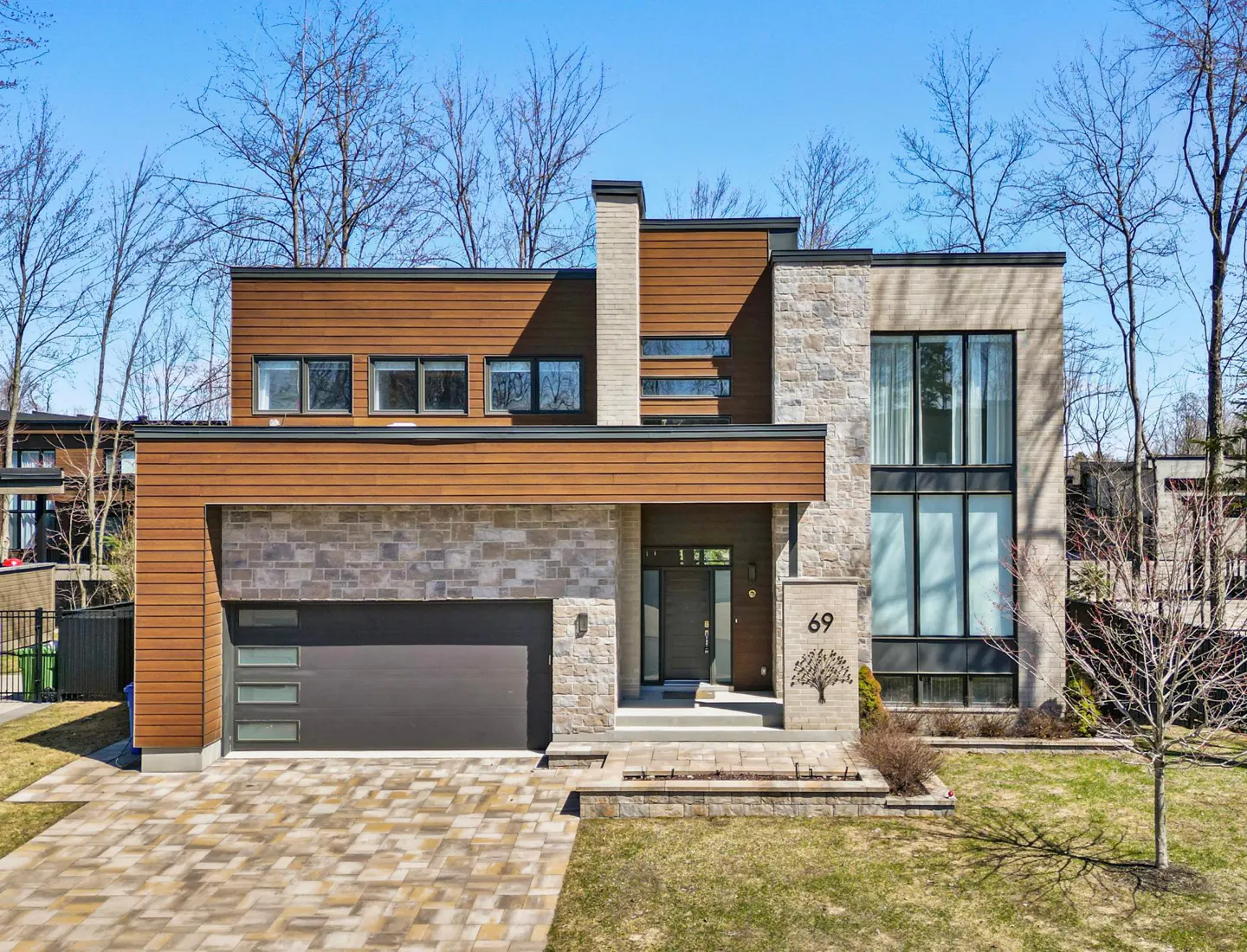 Modern two-story house with wood and stone facade, dark garage door, and number 69 on a stone pillar. Paved driveway and green lawn.