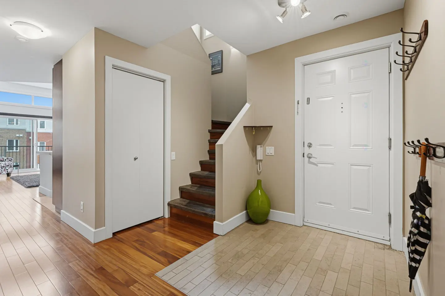 A bright foyer with hardwood floors, a white front door, and a staircase leading upstairs. A green vase sits near the stairs.