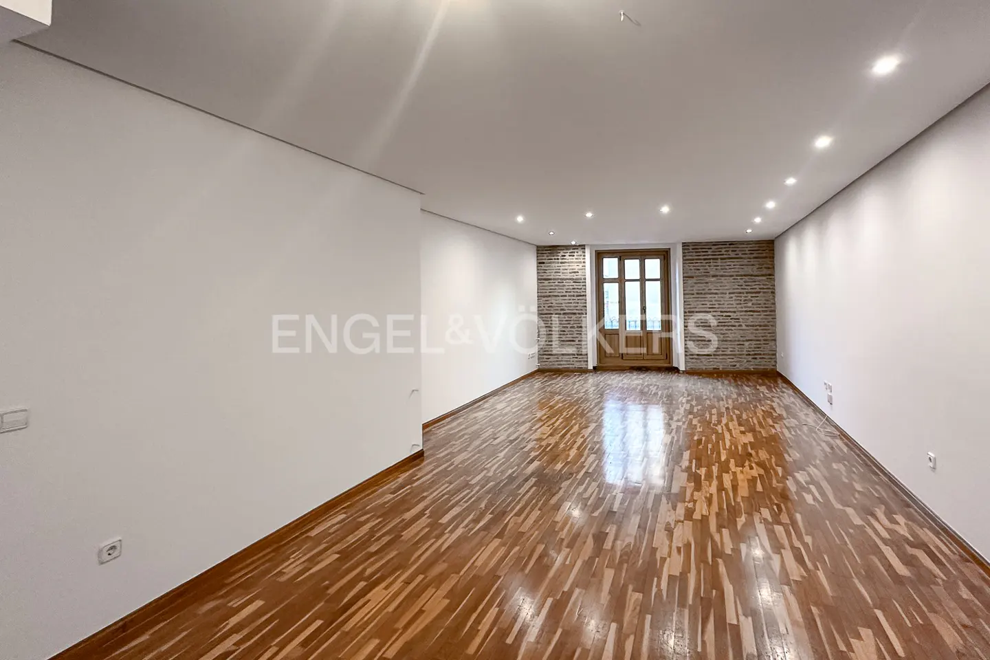 Empty room with white walls, wood floors, and a brick accent wall with a wood-framed window. Engel & Völkers logo on the left.