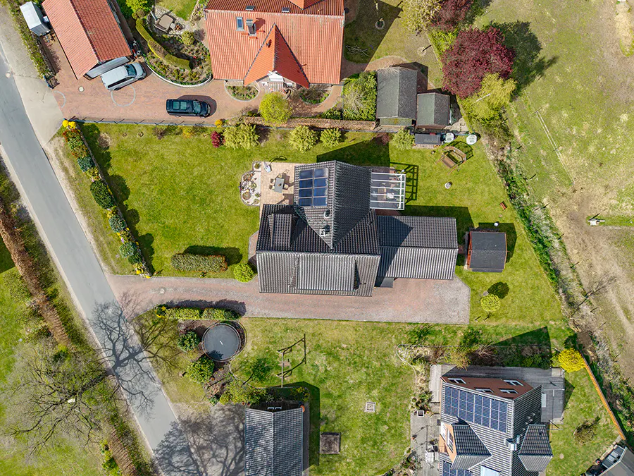 Aerial view of a house with a gray roof and solar panels, surrounded by a green lawn and other houses.
