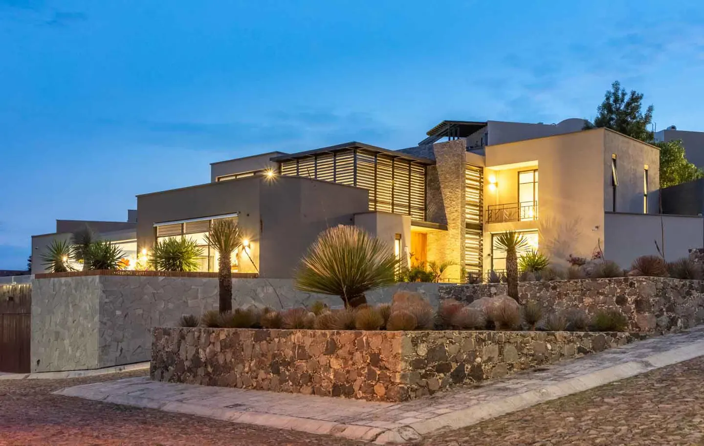 Modern home exterior at dusk. The multi-level house has a stone retaining wall and desert landscaping.