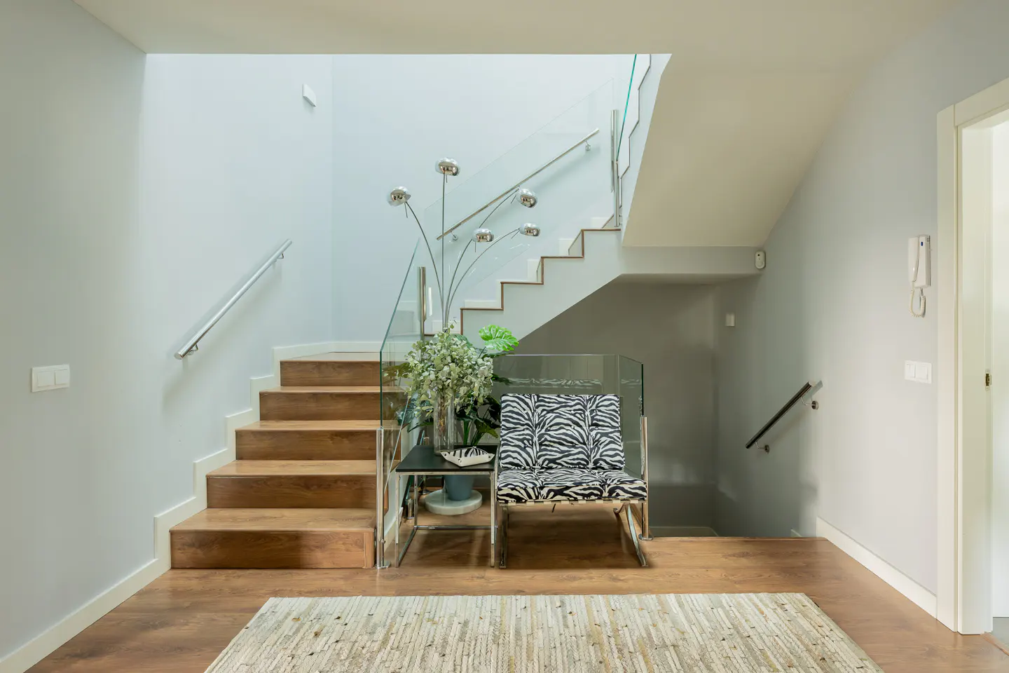 Entryway with wood floors, stairs with glass railings, and a zebra-print chair. A modern floor lamp stands near the stairs.
