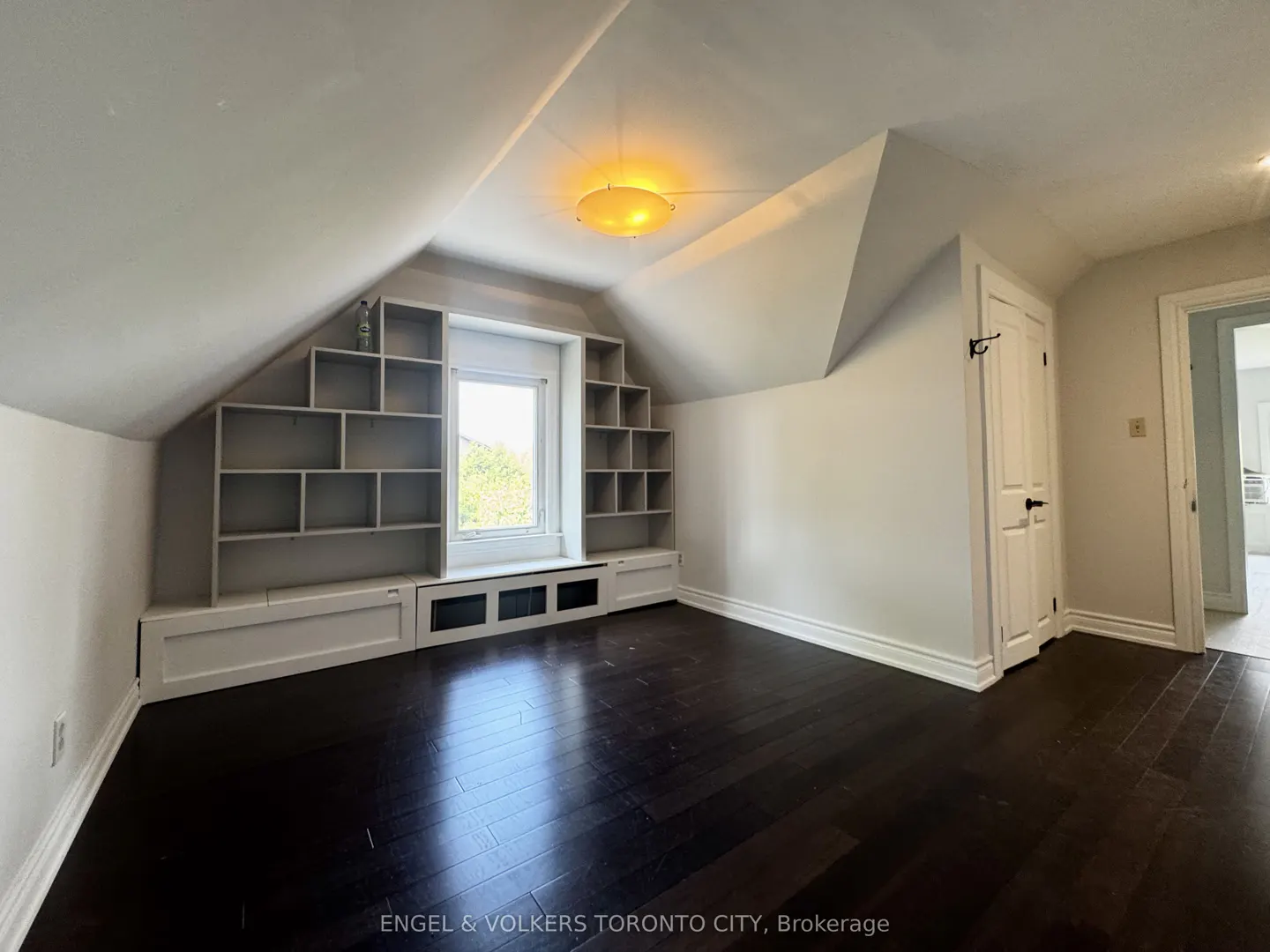 Attic room with dark wood floors, white walls, and built-in shelving around a window. A white door is on the right.