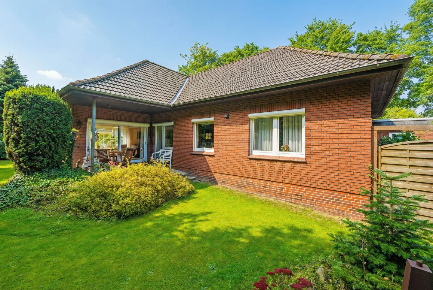 Exterior view of a one-story brick house with a brown tiled roof, green lawn, and patio furniture visible through sliding glass doors.