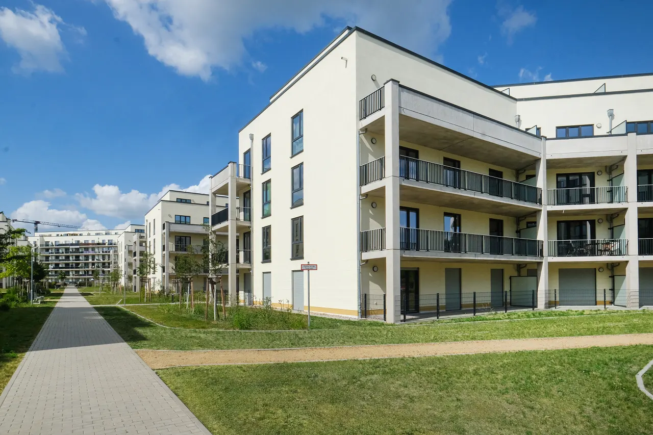 Modern apartment buildings with balconies line a walkway and green space under a blue sky.