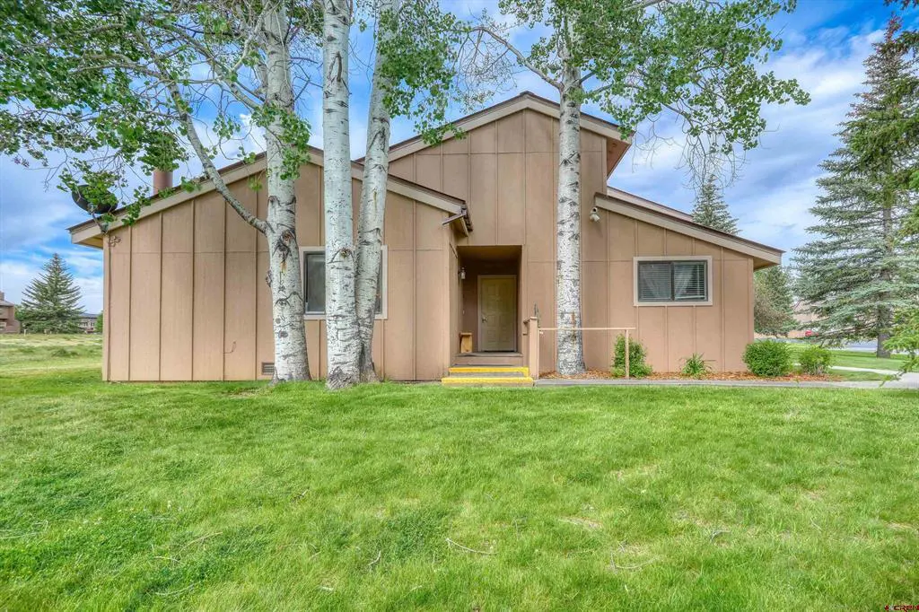 Tan-colored house with vertical siding, front entrance, and tall aspen trees in a grassy yard under a blue sky.