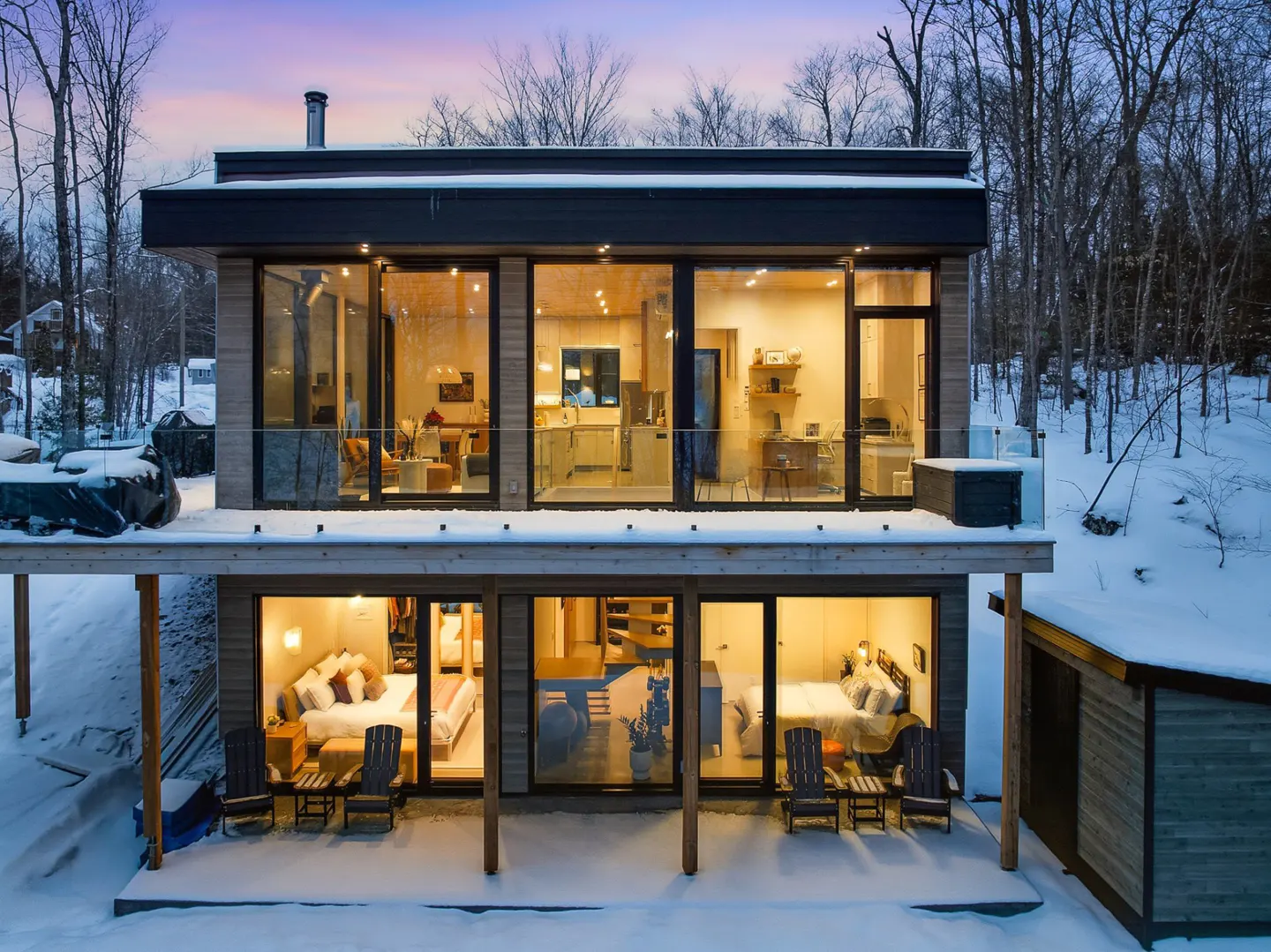 Two-story modern home with large windows, revealing interior spaces. Snow covers the ground and roof. Trees in the background.
