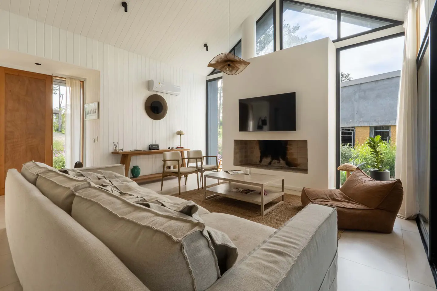 Bright living room with white walls, large windows, and a beige sofa. A TV is mounted above a fireplace. A wooden table and chairs sit near a sliding door.
