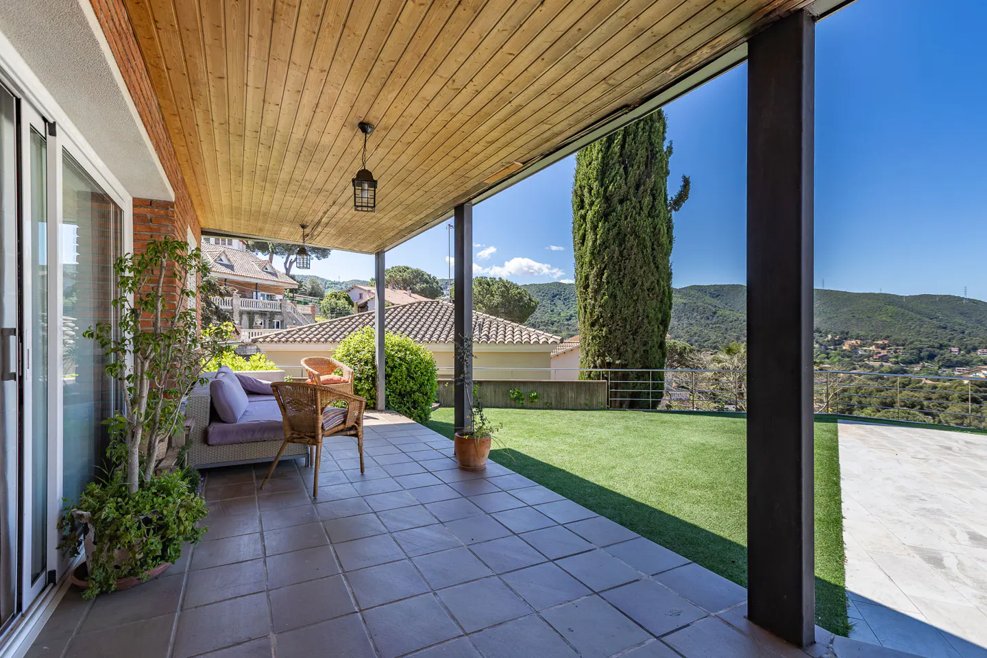 Covered patio with wicker furniture, tile floor, and wood ceiling. Green lawn and trees in the background.