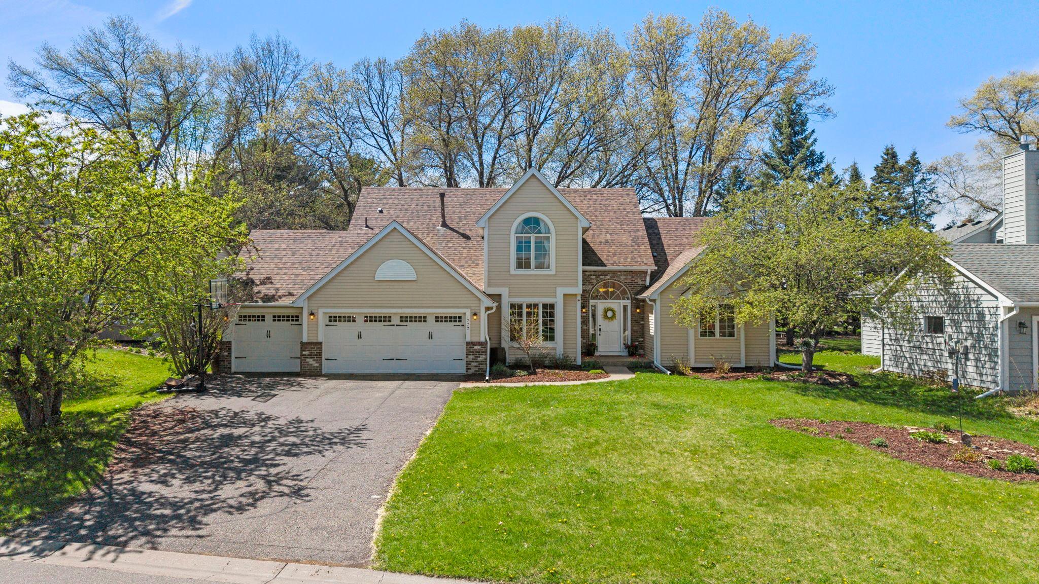 Beige two-story house with a brown roof, white garage doors, and a green lawn on a sunny day.