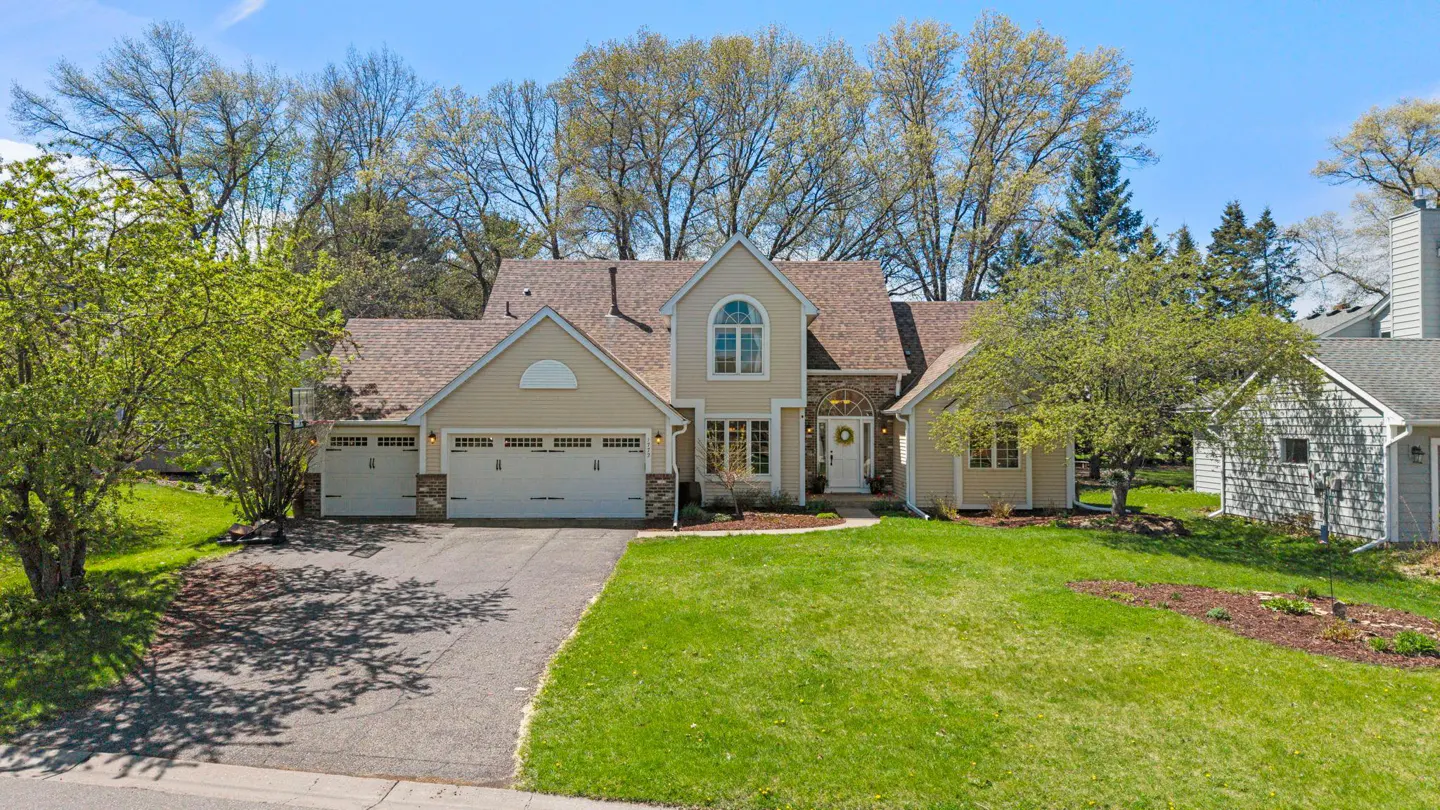 Beige two-story house with a brown roof, white garage doors, and a green lawn on a sunny day.