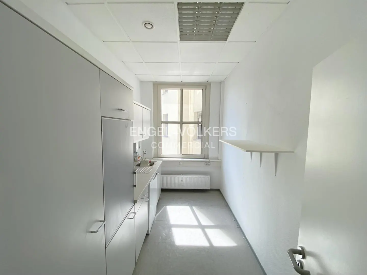 Bright, empty office kitchenette with white cabinets, sink, and a window. A shelf is mounted on the right wall.