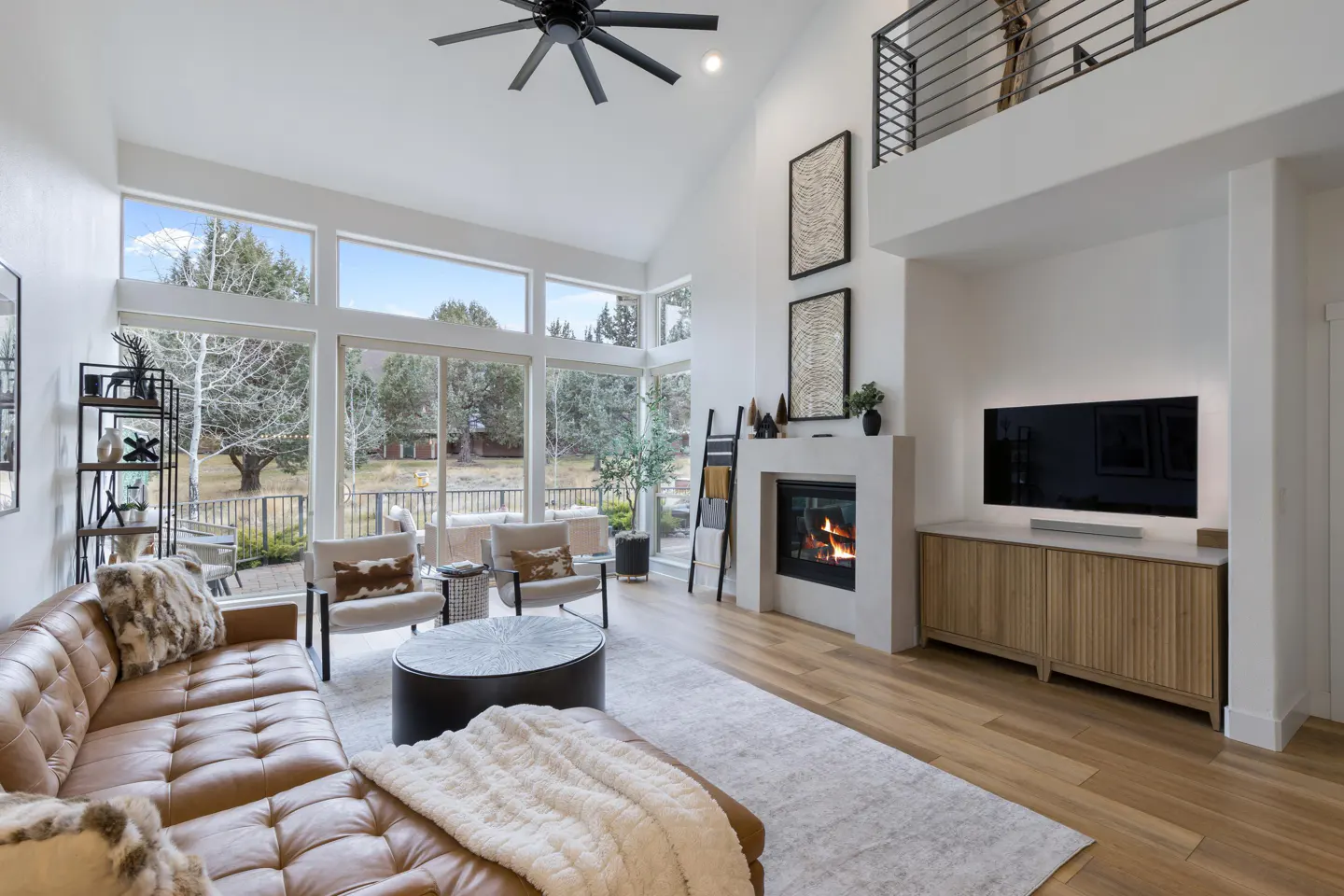 Bright living room with a brown leather sofa, fireplace, and large windows overlooking trees. A black ceiling fan hangs above.